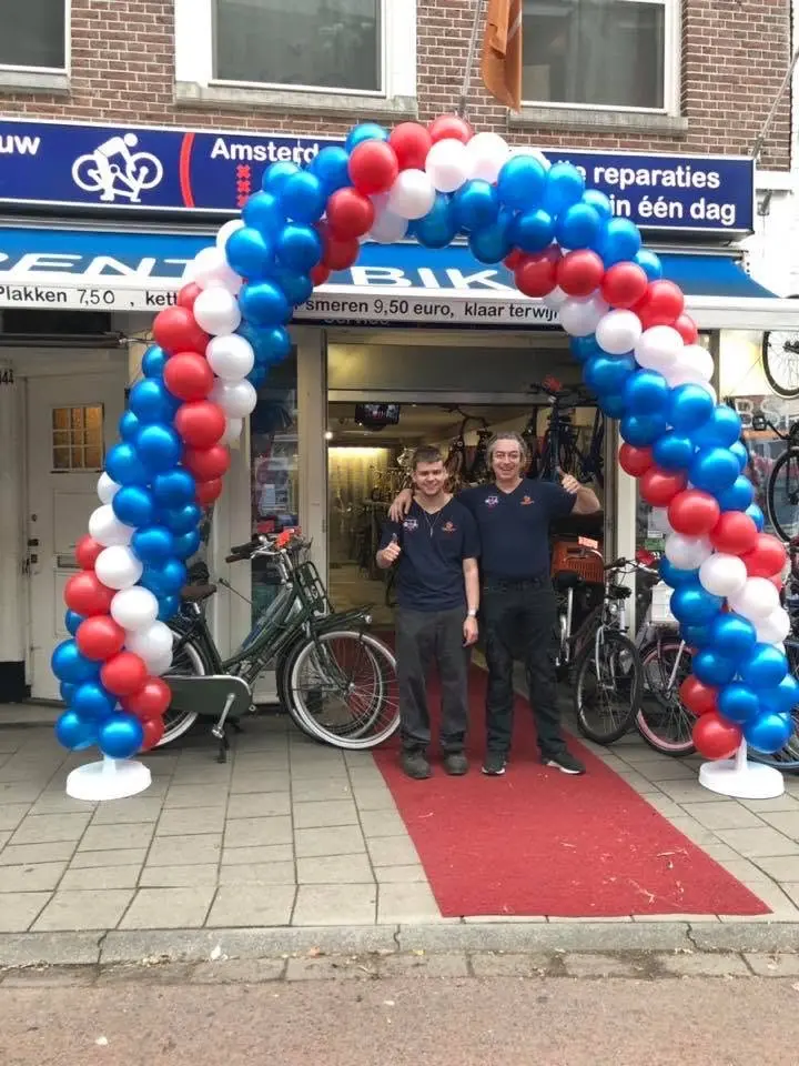 Twee mannen voor de deur van de winkel, onder een boog van rode, witte en blauwe ballonnen.